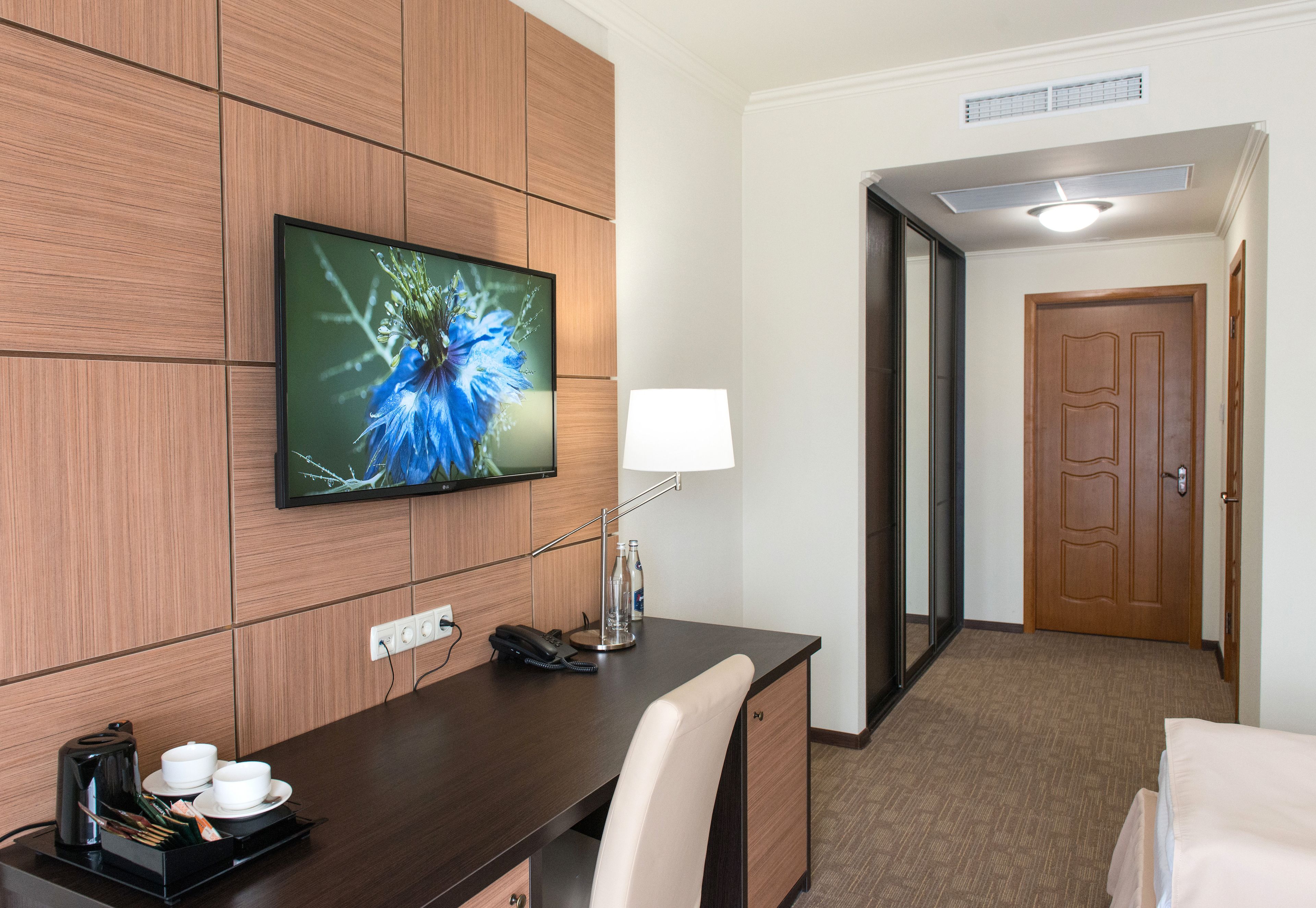 A hotel room with a wooden desk, a television mounted on the wall, a lamp, a telephone, and a door leading to another room.