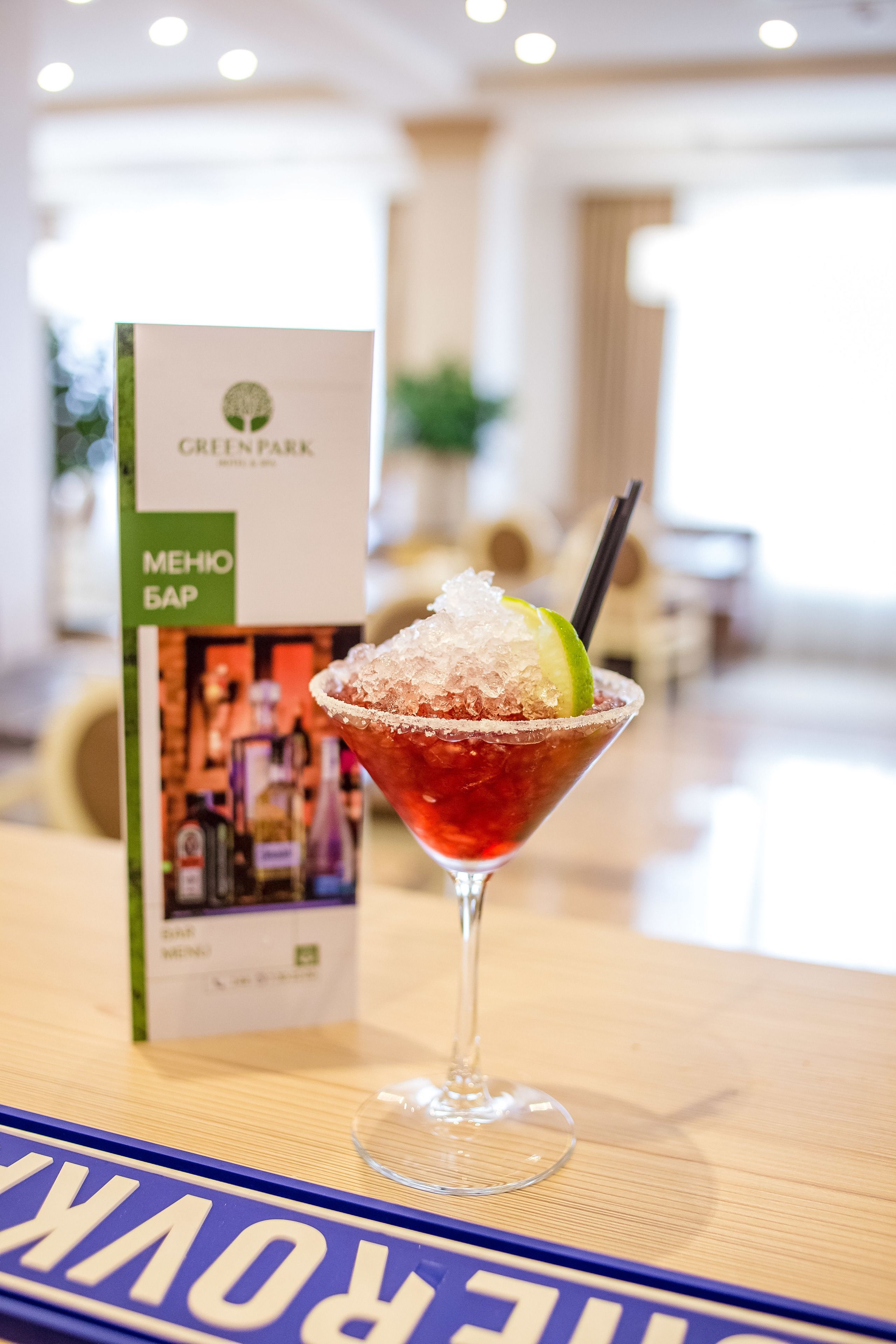 Una copa de martini con una rodaja de lima y hielo sobre una mesa de madera, con un menú de Green Park al fondo.