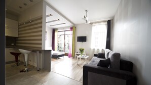 A modern living room with a grey sofa, a small white table, and a TV mounted on the wall. There is a kitchen area with a red bar stool and a white chair. - Rint - Centrum Nowy Świat (Bialystok)