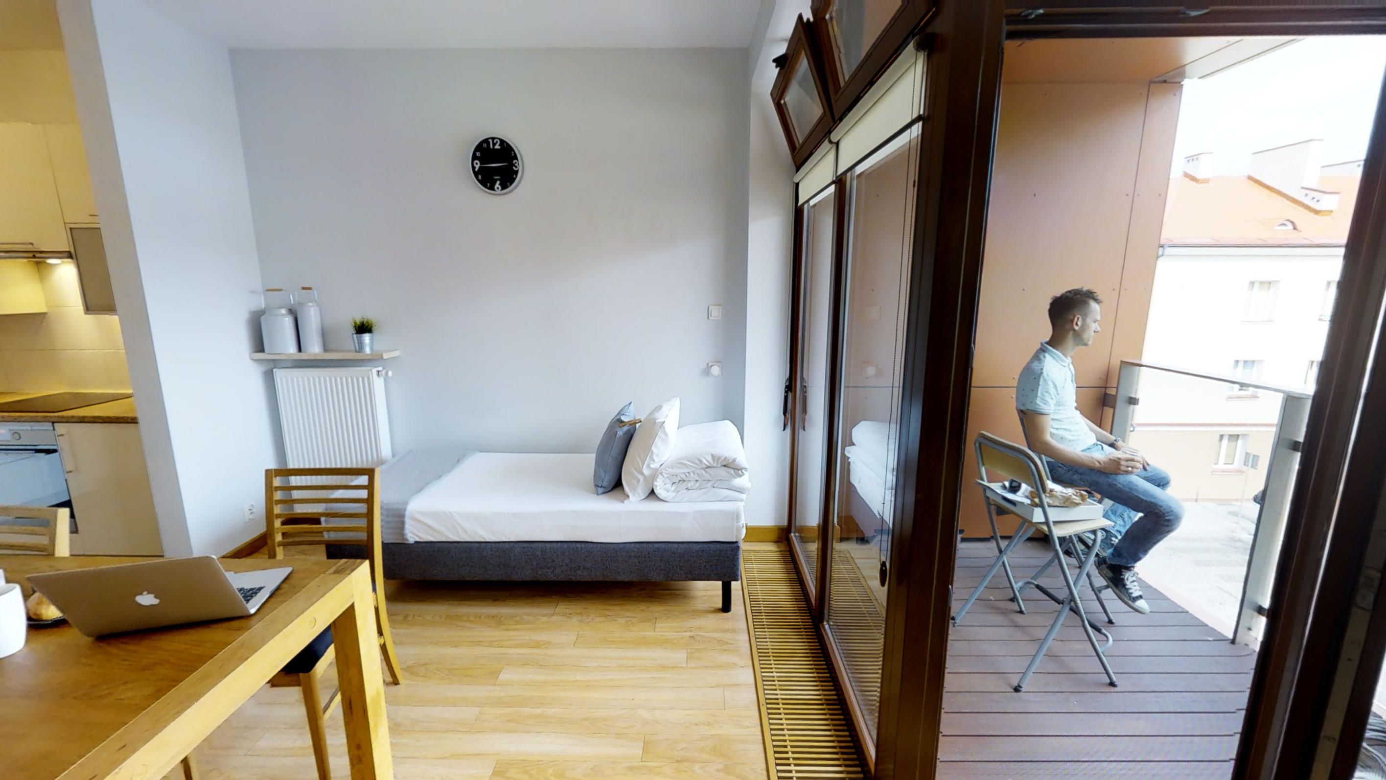 A bedroom with a bed, a desk with a laptop, a radiator, and a window overlooking a balcony.
