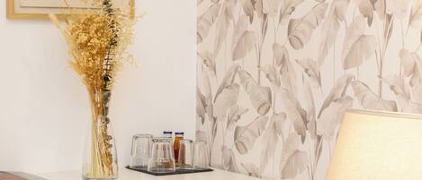 A room with a white desk, a vase with dried flowers, a framed abstract artwork, a wall with a leaf pattern, and a lamp.