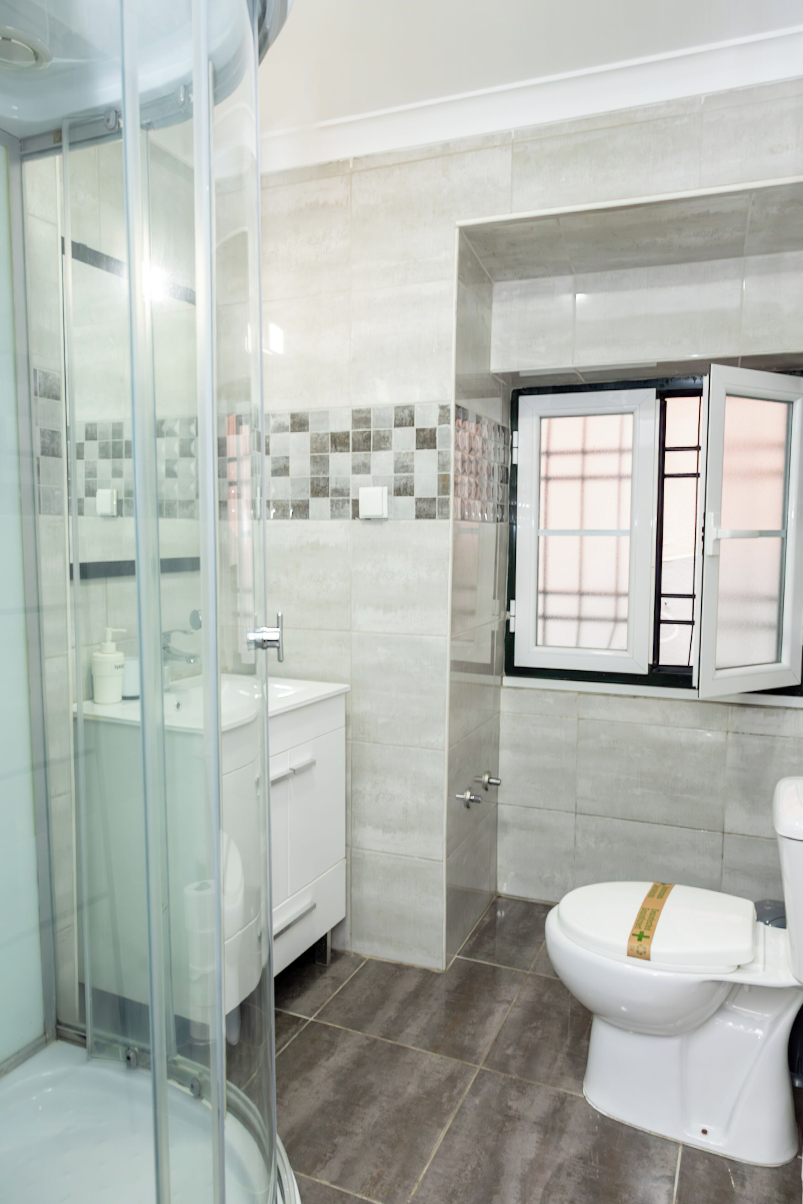 A modern bathroom with a glass shower enclosure, a white sink, and a toilet with a window above it.
