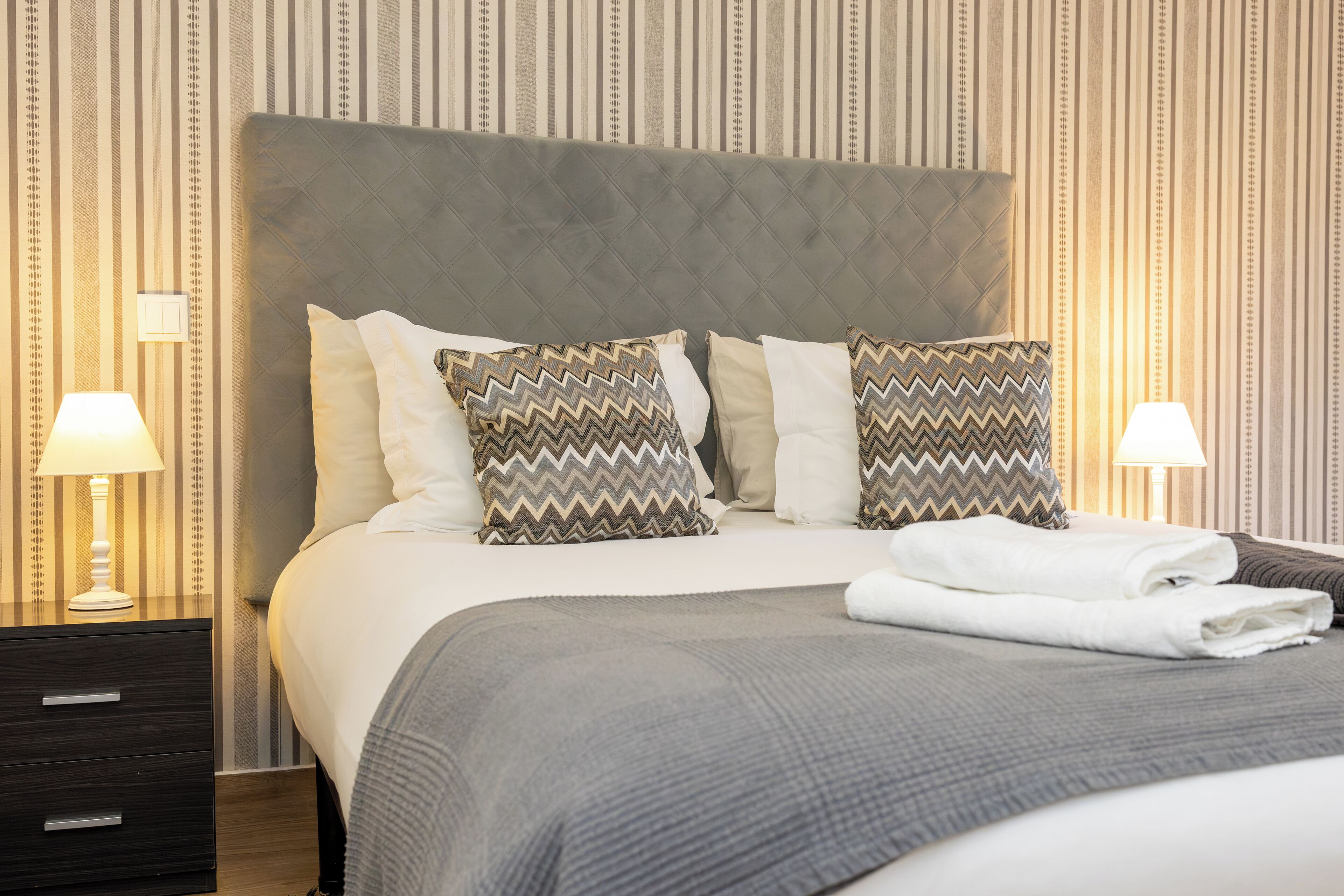 A neatly made bed with a grey headboard, two pillows with zigzag patterns, and a folded white towel on the bed.
