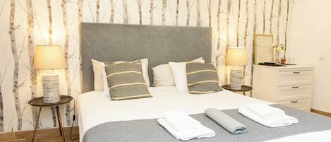Una cama bien hecha con almohadas, una mesita de noche con lámpara y un aparador con espejo.