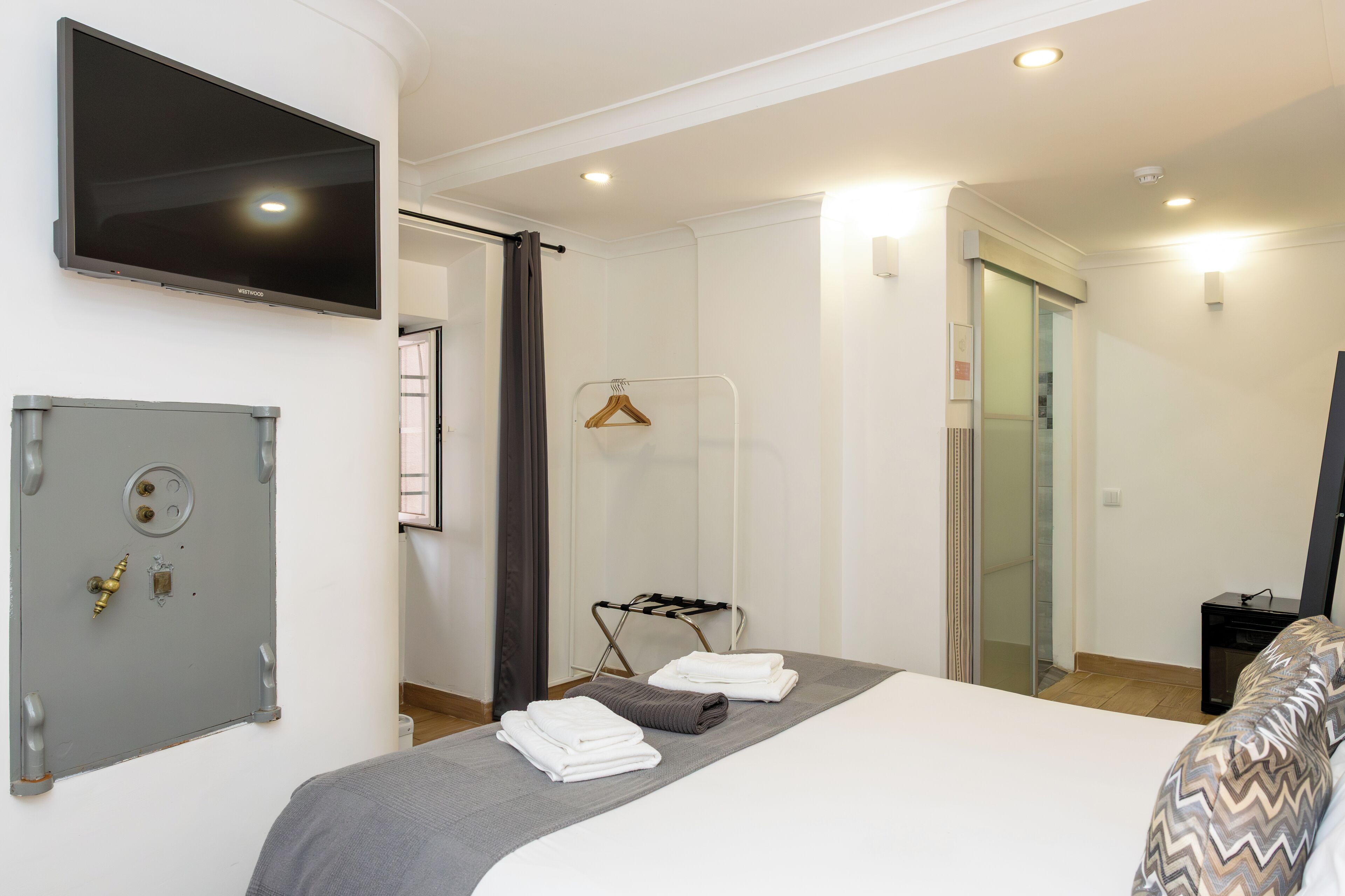 A hotel room with a bed, a TV mounted on the wall, a safe, and a shower area.