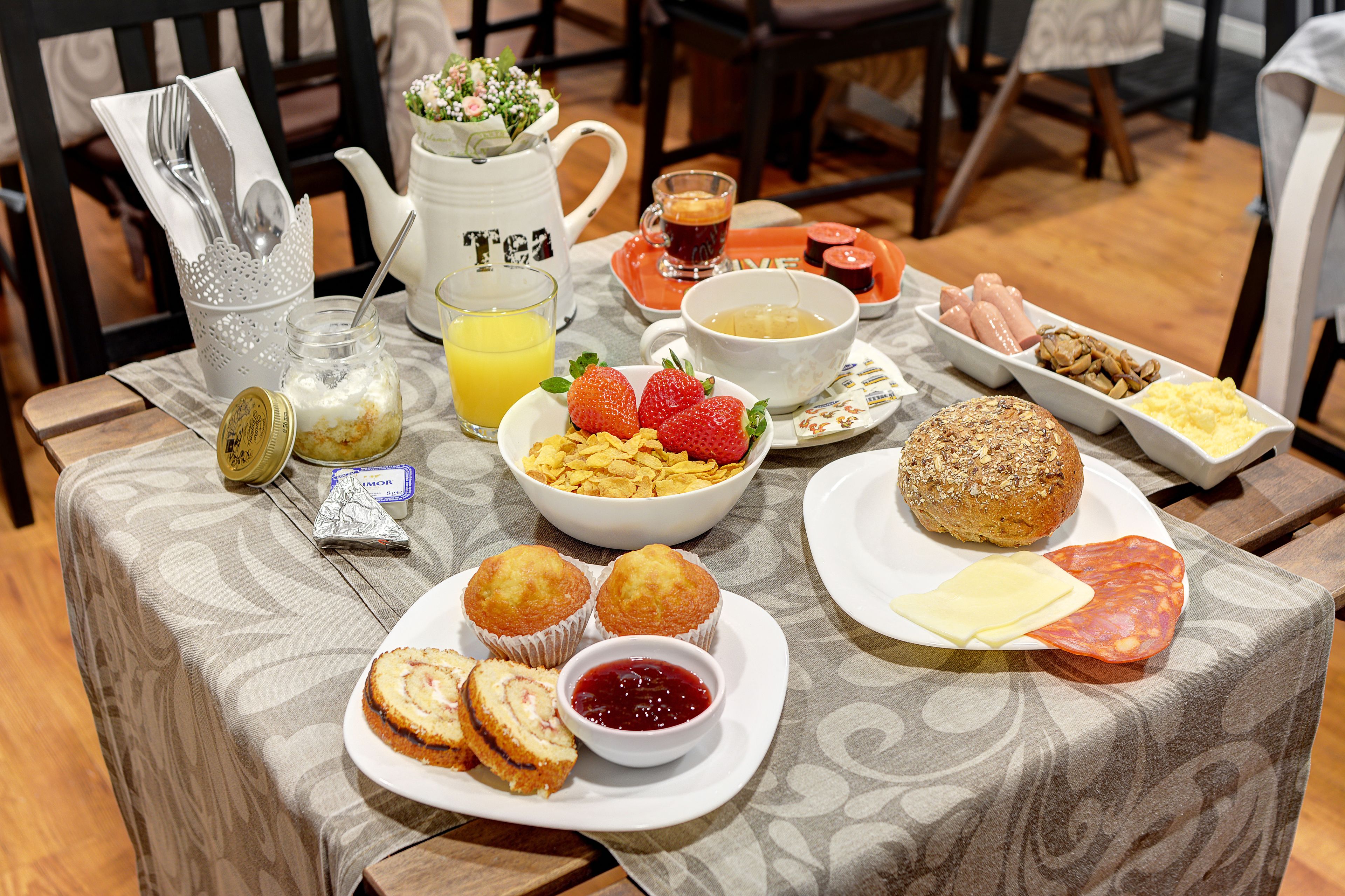 A breakfast table with various food items, a pitcher, a glass of orange juice, and a teapot.