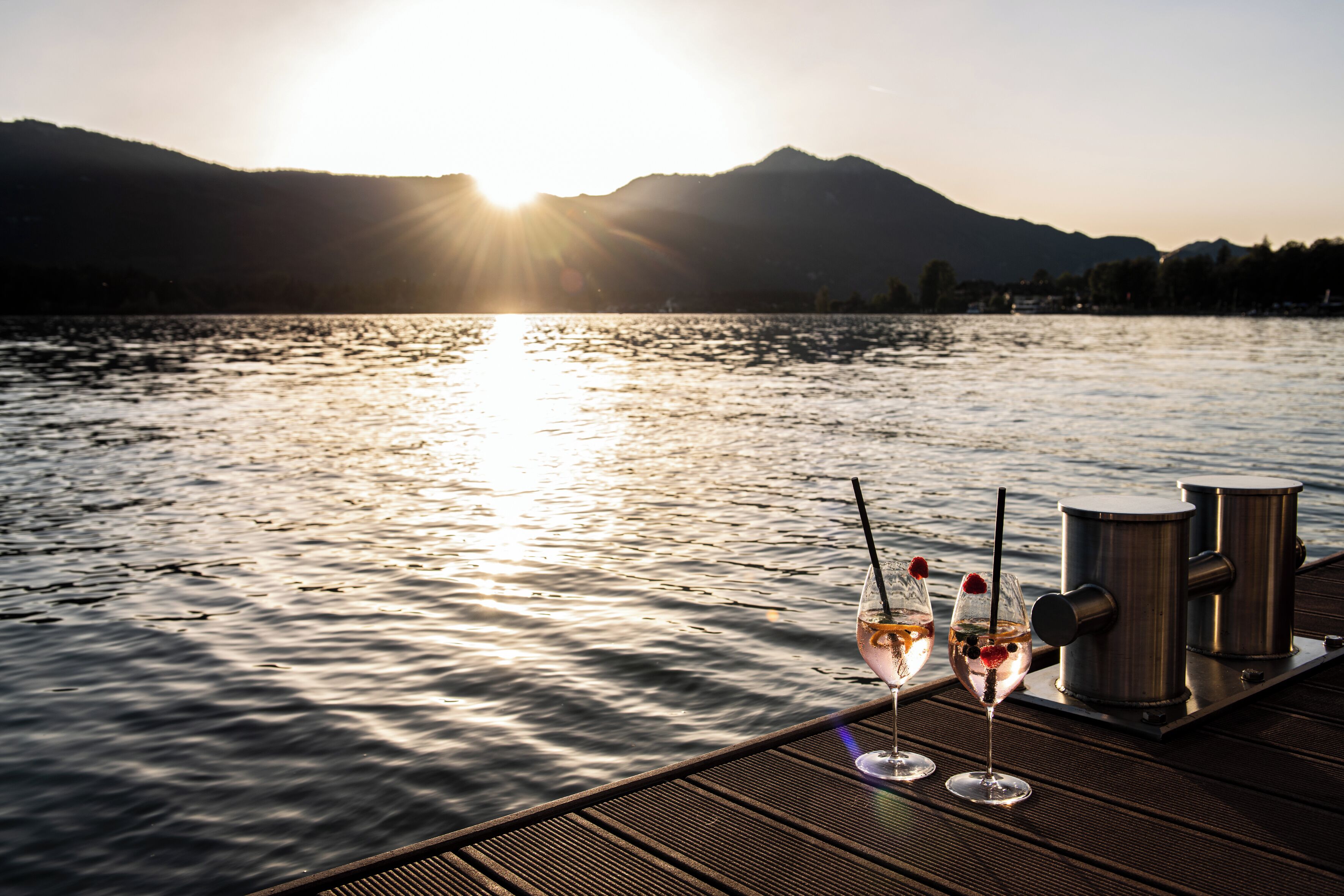 Muellé de madera con dos copas de vino y una cafetera, con vista a un lago tranquilo y montañas al fondo.