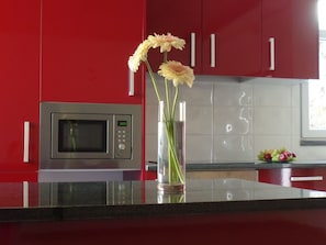 A kitchen with red cabinets, a microwave, a vase with flowers, and a fruit bowl. - Apartamentos Príncipe Apart-Center (Horta)