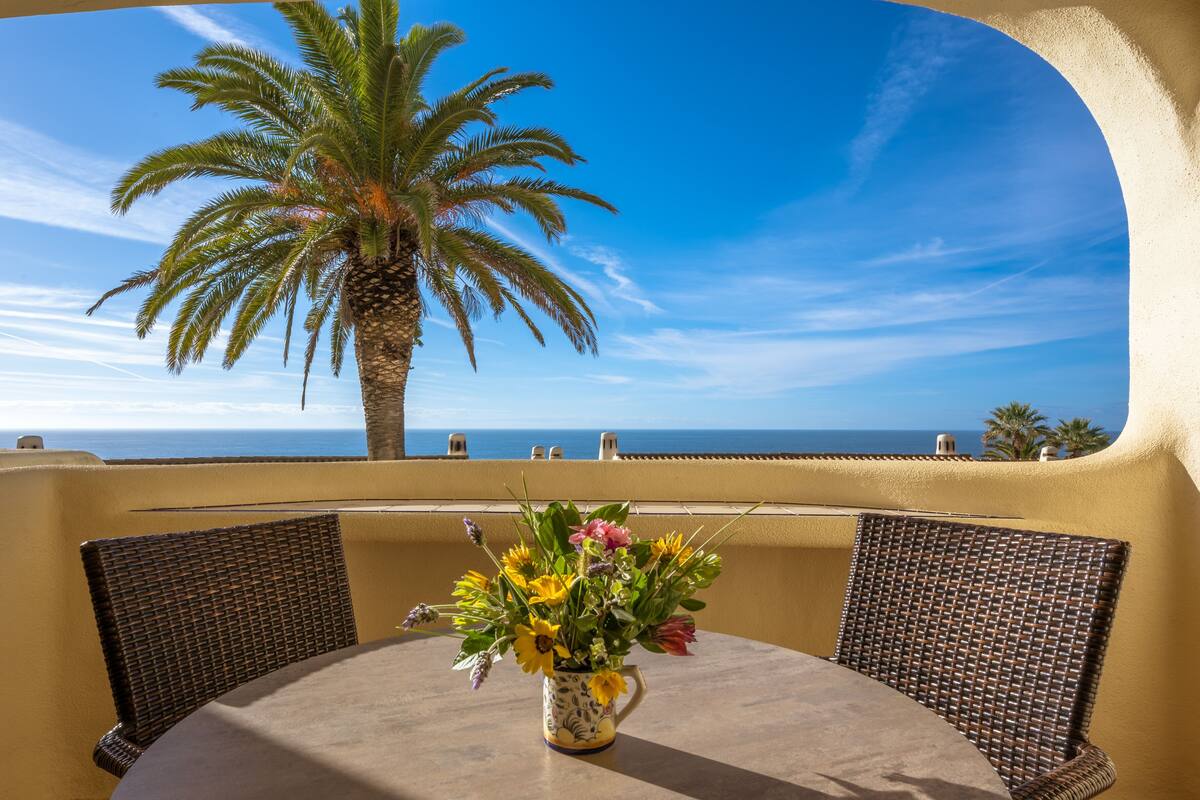 A round table with a vase of flowers, two wicker chairs, and a view of the ocean and palm trees.