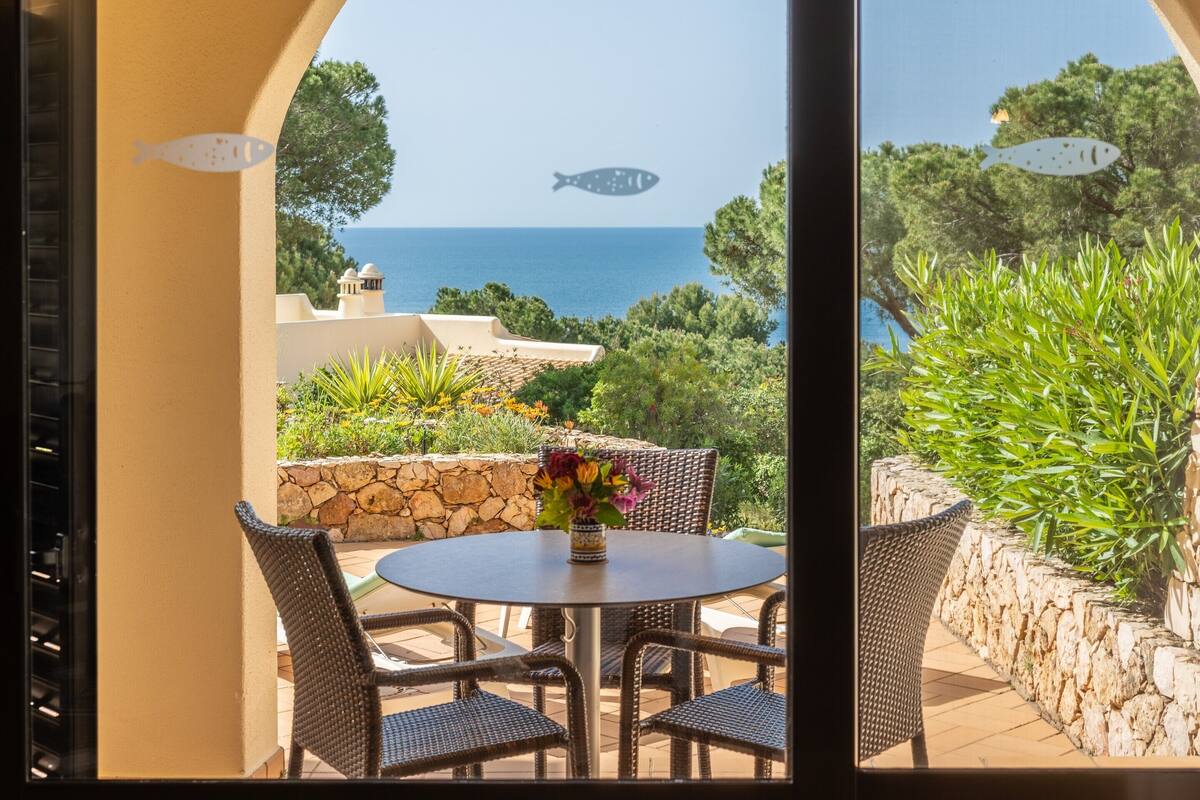 A patio with wicker furniture and a table with a vase of flowers, overlooking a scenic view of the sea and greenery.