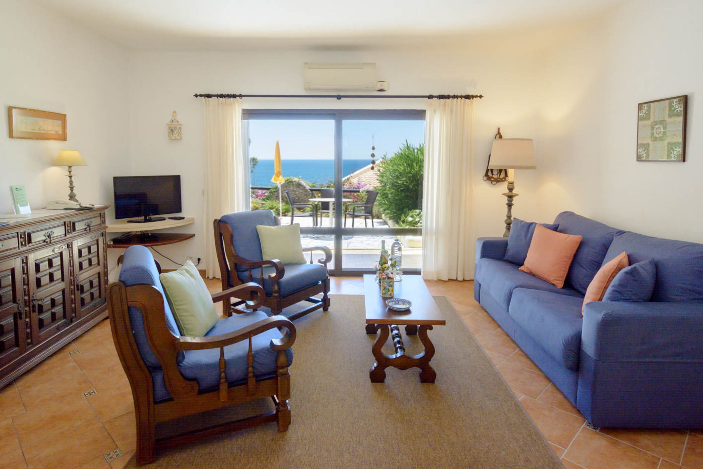 A living room with a sofa, two armchairs, a coffee table, a TV, and a view of the sea through large windows.