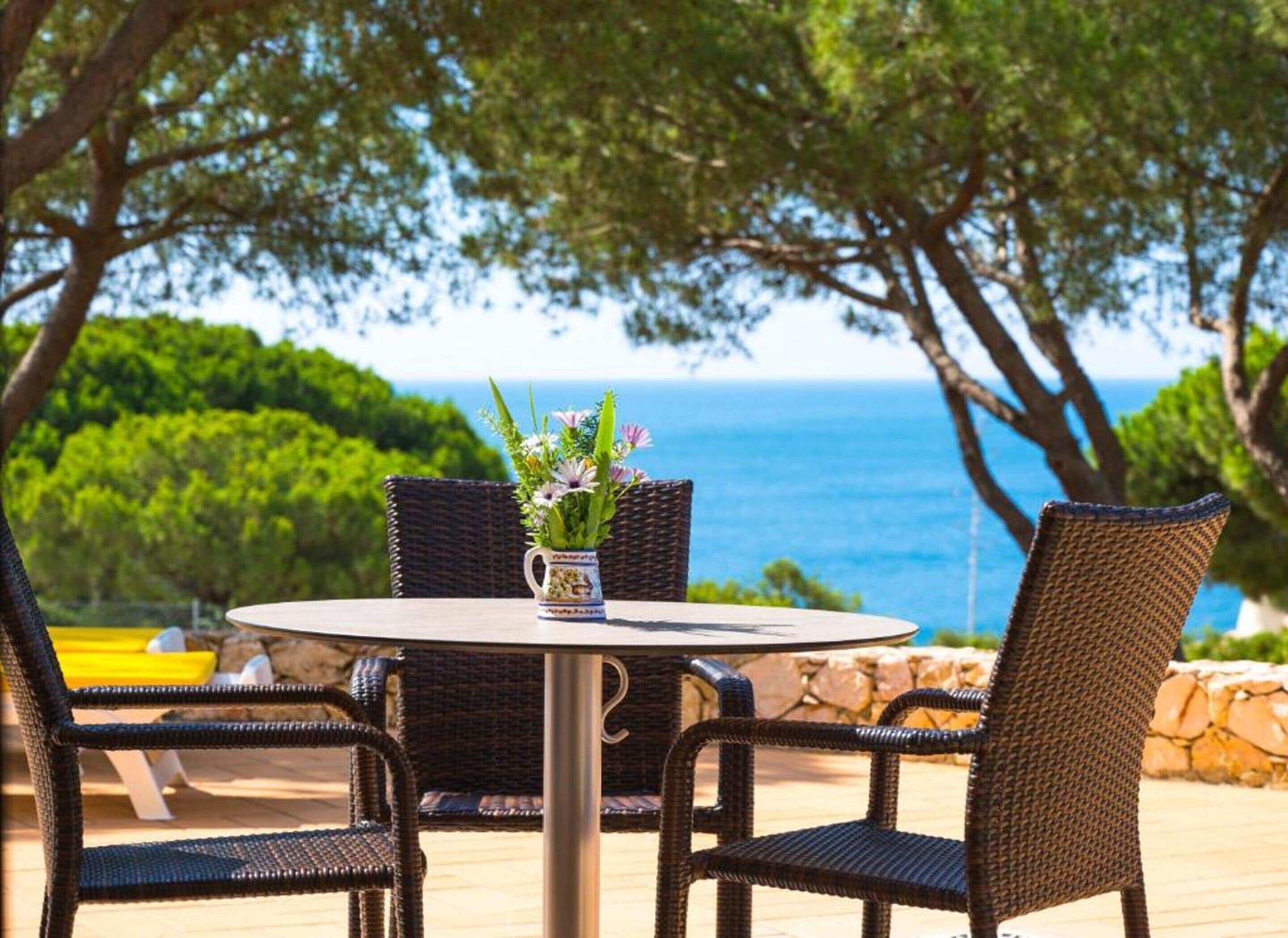 Una mesa redonda con sillas de mimbre, un florero con flores y vista al mar.