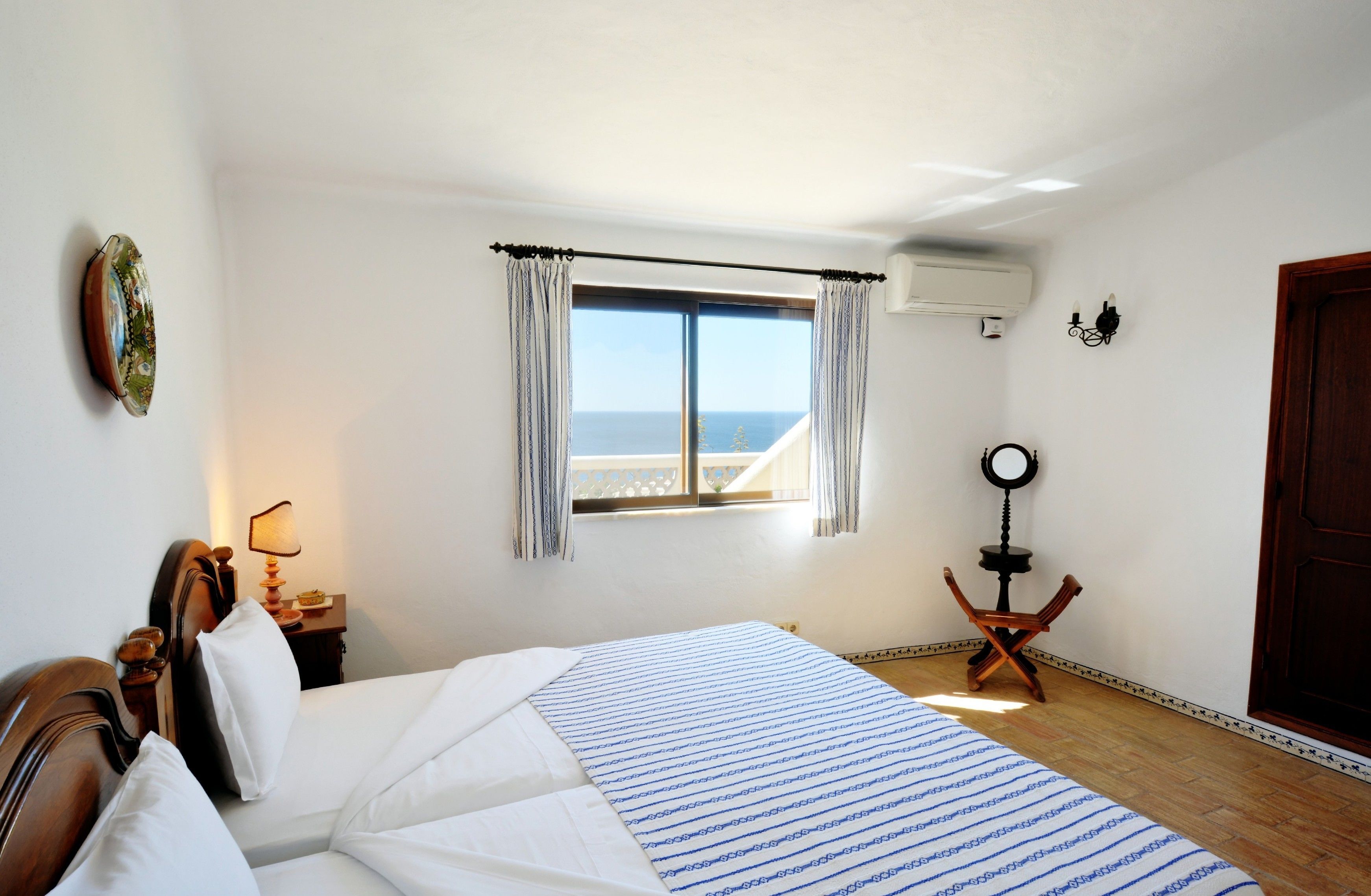 Un dormitorio con cama, ventana con cortinas, lámpara de noche, una mesita y vista al mar.