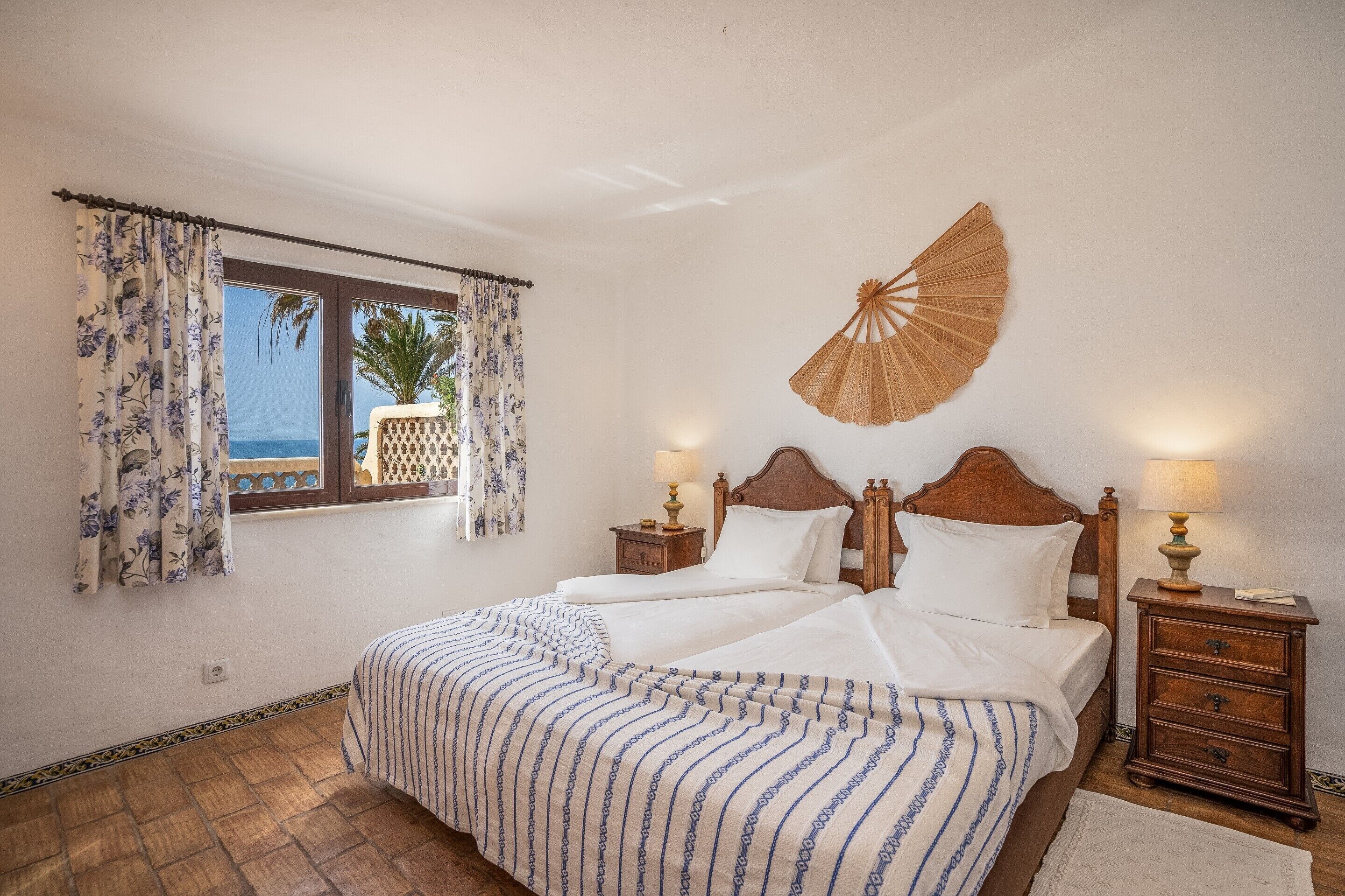 A bedroom with a large bed, two bedside tables with lamps, a decorative fan, and a window with curtains.