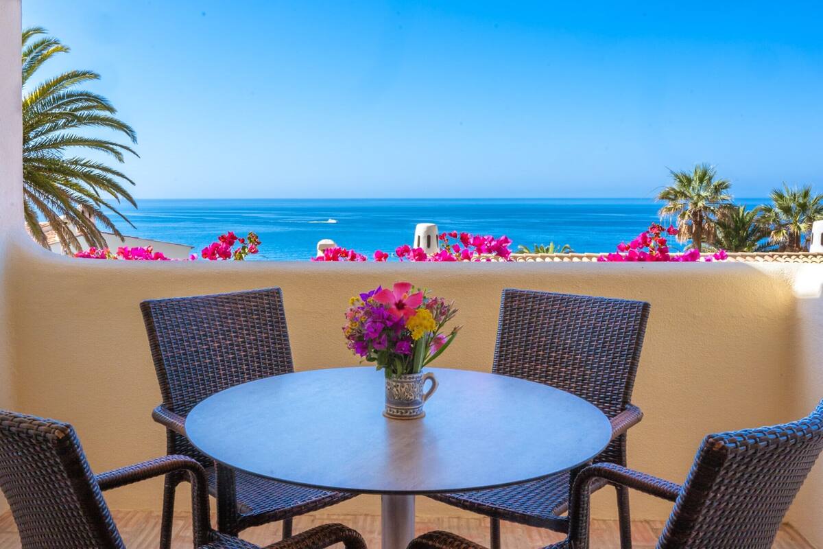 A round table with wicker chairs and a vase of flowers, overlooking a beach with palm trees and the ocean.