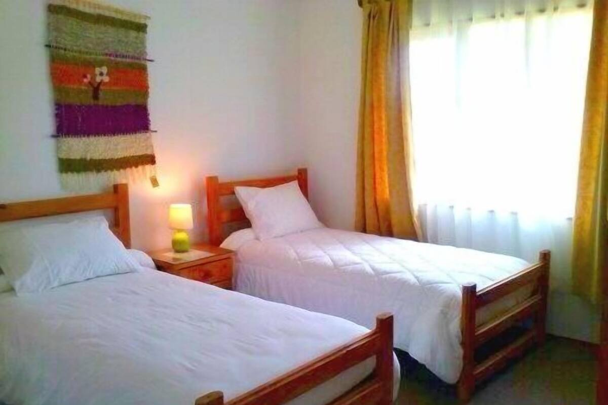Una habitación con dos camas individuales, cabeceros de madera, ropa de cama blanca, una mesita de noche con una lámpara y un tapiz colorido en la pared.