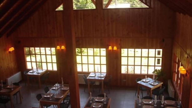 Un comedor rústico de madera con techos altos y ventanas amplias. Las mesas están preparadas con platos y cubiertos.