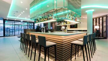 Un espace bar moderne doté d’un îlot central et de nombreux tabourets, d’un plafond haut avec des luminaires encastrés et d’une vue sur l’extérieur grâce à de grandes fenêtres.