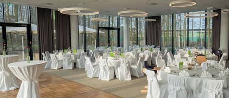 A banquet hall with round tables, white tablecloths, and chairs, large windows, and modern circular ceiling lights.