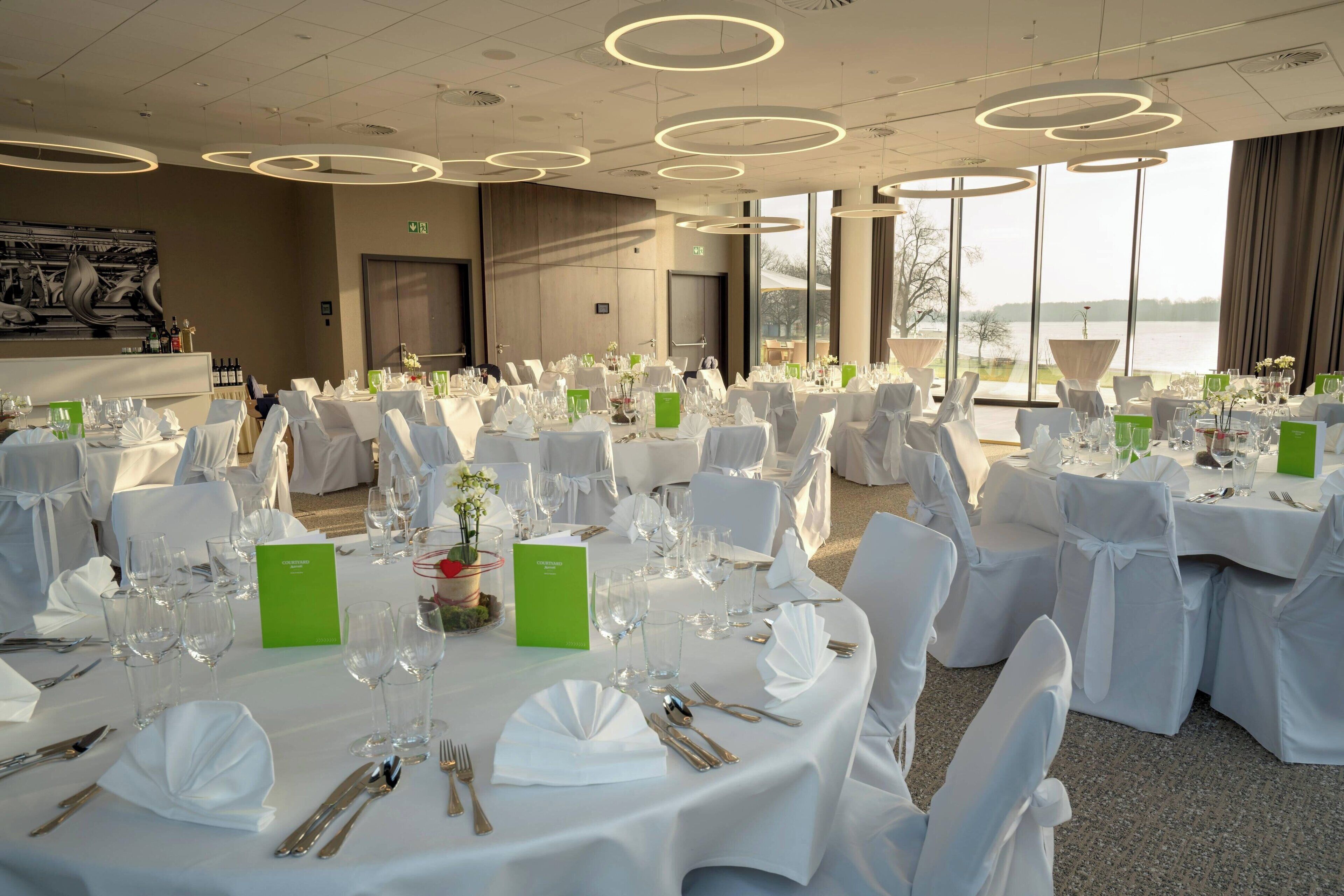 Une salle de banquet avec des tables rondes disposées pour un événement formel, de grandes fenêtres offrant une vue sur l’extérieur et un design moderne au niveau du plafond.