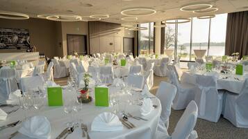 Une salle de banquet avec des tables rondes disposées pour un événement formel, de grandes fenêtres offrant une vue sur l’extérieur et un design moderne au niveau du plafond.