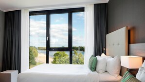 A hotel room with a large window, a bed, a bedside table, and a lamp. - Courtyard by Marriott Wolfsburg (Wolfsburg)