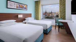 Habitación de hotel con dos camas, un escritorio, una silla, un televisor y ventana con vista a la ciudad.