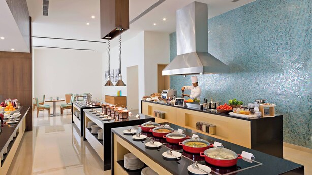 Un comedor moderno de hotel con mostradores de buffet, arreglos de asientos y un chef preparando comida.