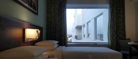 A hotel room with two beds, a nightstand with a lamp, a view of a cityscape through large windows, and a painting on the wall.