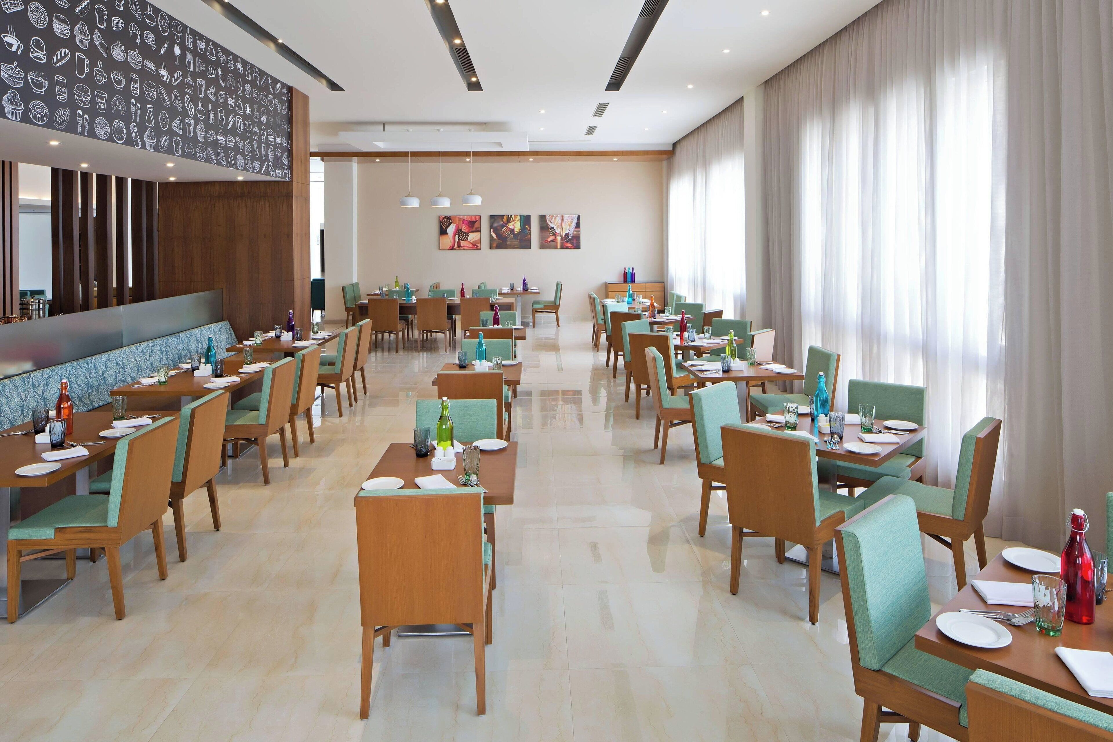Un amplio salón comedor de hotel con varias mesas y sillas, una larga barra y un gran ventanal con cortinas.