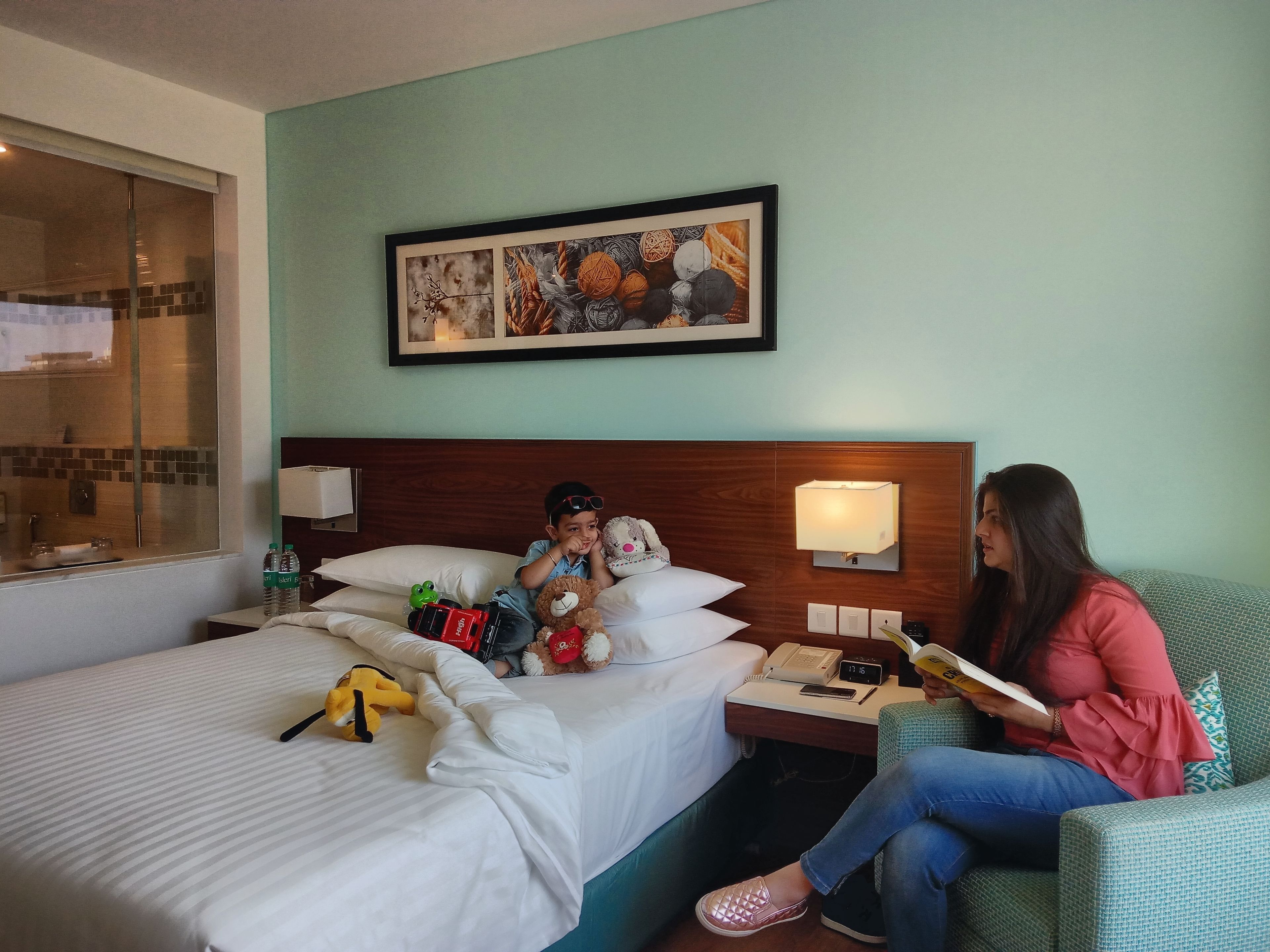 A hotel room with a bed, a nightstand with a lamp, a framed picture on the wall, and a window with a view of the outside.