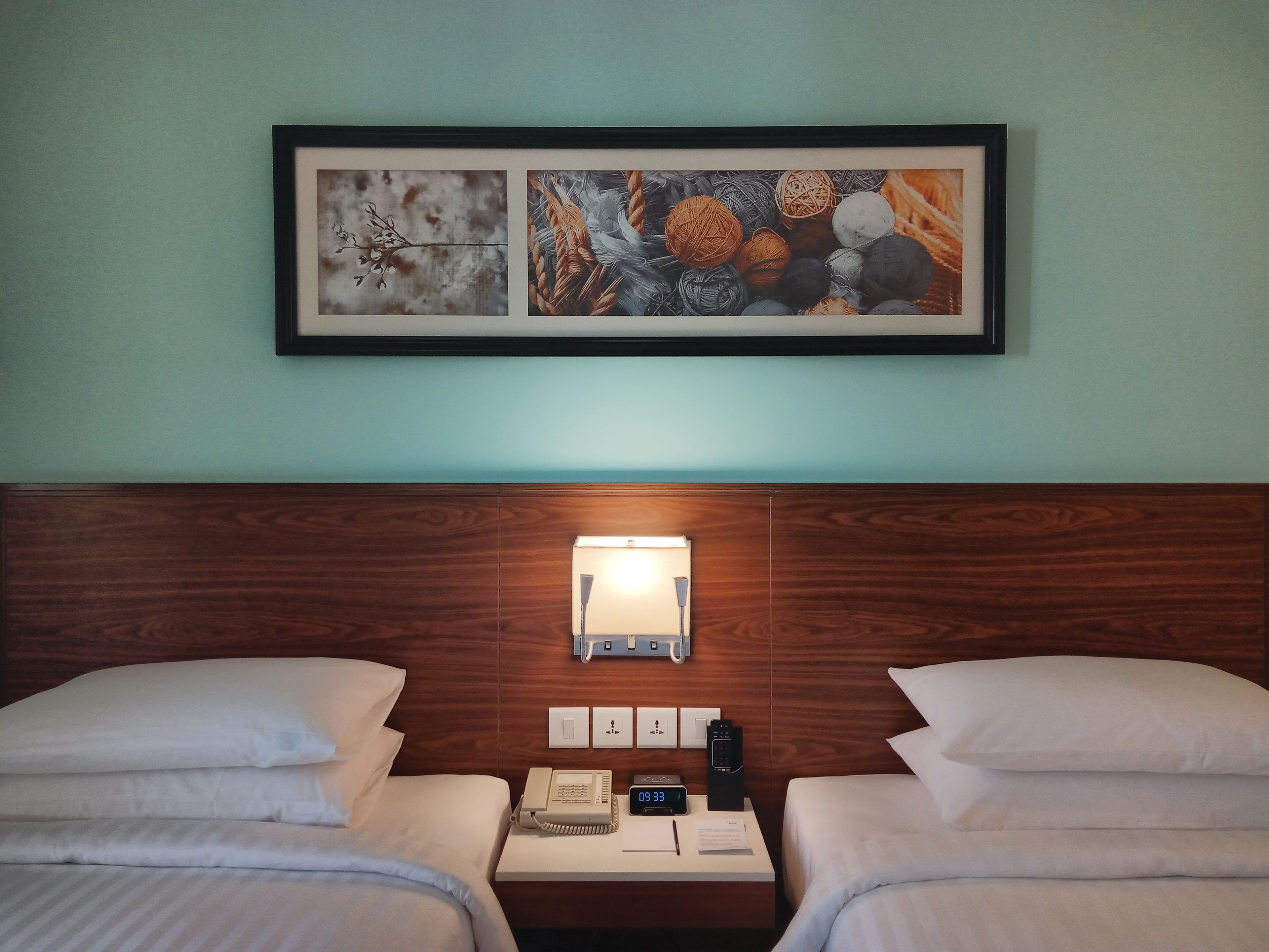A hotel room with two beds, a nightstand with a phone, a clock, and a wall-mounted light.
