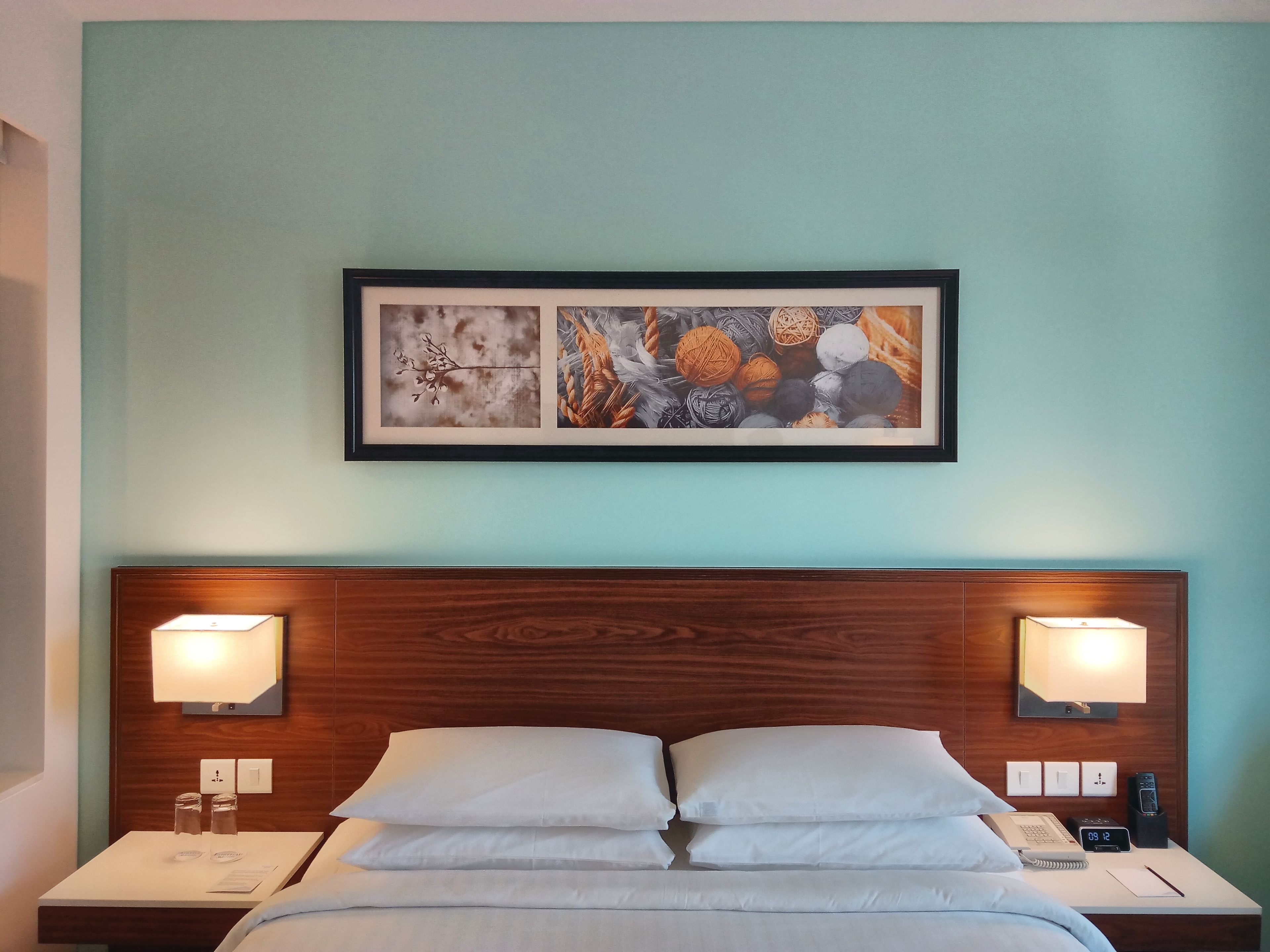 Een hotelkamer met een bed, twee bedlampen, een nachtkastje met een telefoon en een lamp, en een ingelijst kunstwerk boven het bed.