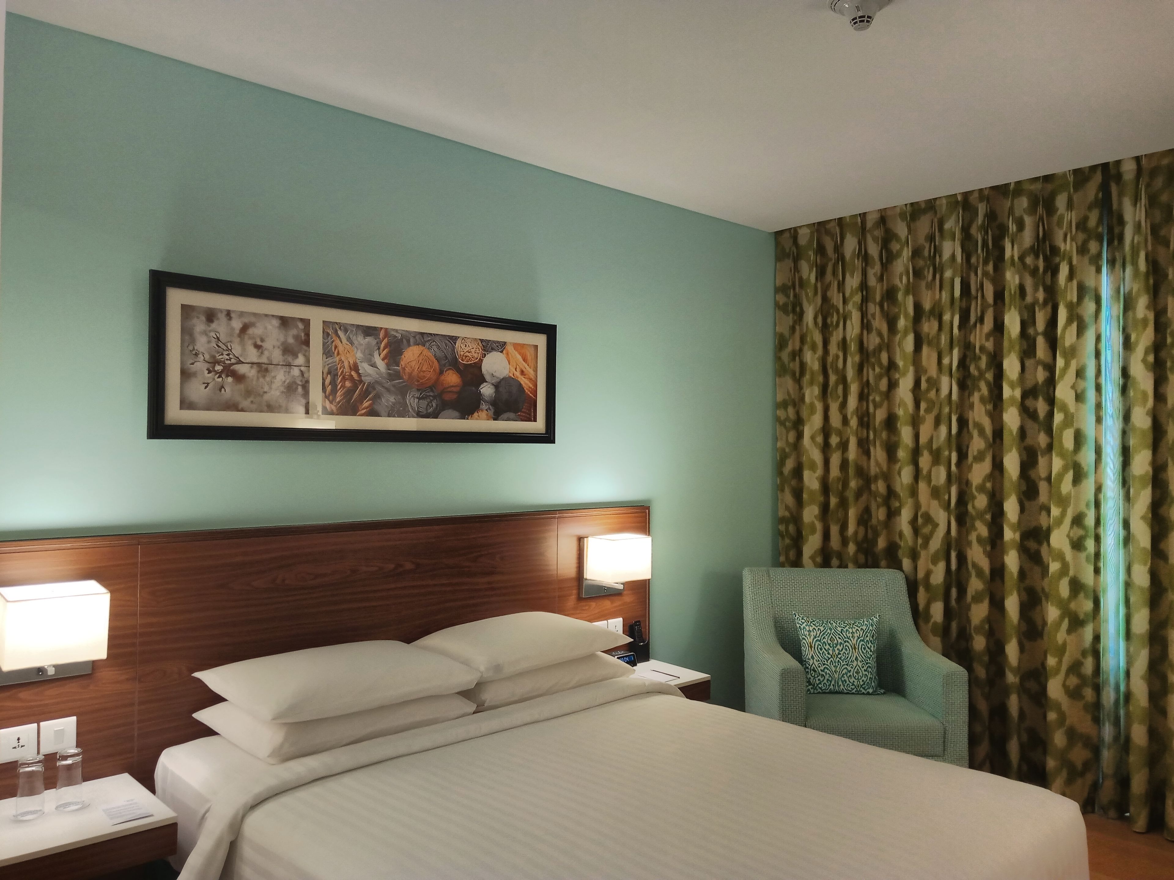 A hotel room with a bed, a nightstand, a lamp, a chair, and a patterned curtain.