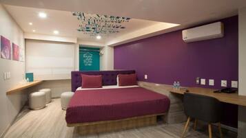 A modern hotel room with a large bed, a desk, and a chair. There is a ceiling light fixture and an air conditioning unit on the wall.