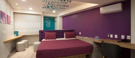 A modern hotel room with a large bed, a desk, and a chair. There is a ceiling light fixture and an air conditioning unit on the wall.