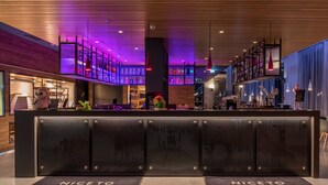 Un espace d’accueil d’hôtel au design moderne, comprenant un comptoir de réception central, un bar avec des étagères remplies de bouteilles et une statue de hibou décorative.