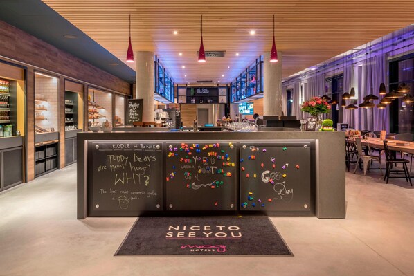 A modern hotel lobby with a central reception desk, a bar area, and seating arrangements. - Moxy Munich Messe (Aschheim)