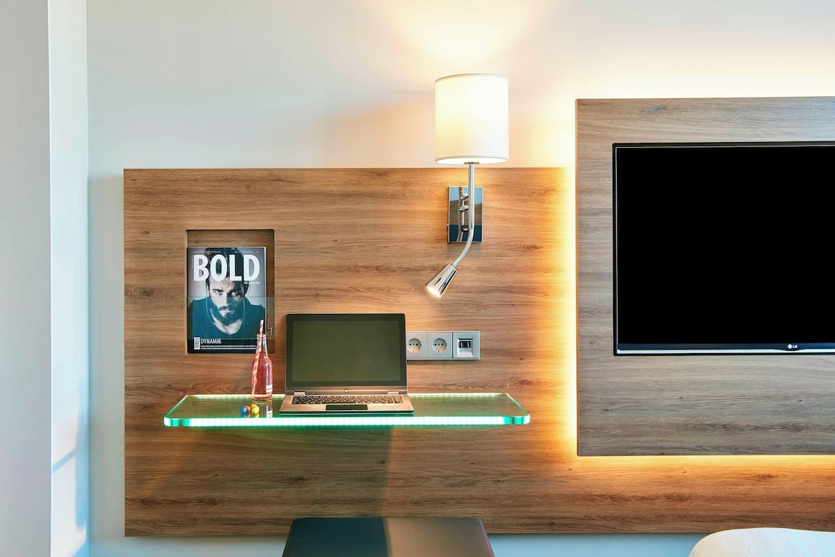 Ein modernes Zimmer mit einer Holzwand, einem Glastischchen mit Laptop, Lampe und einem Poster.