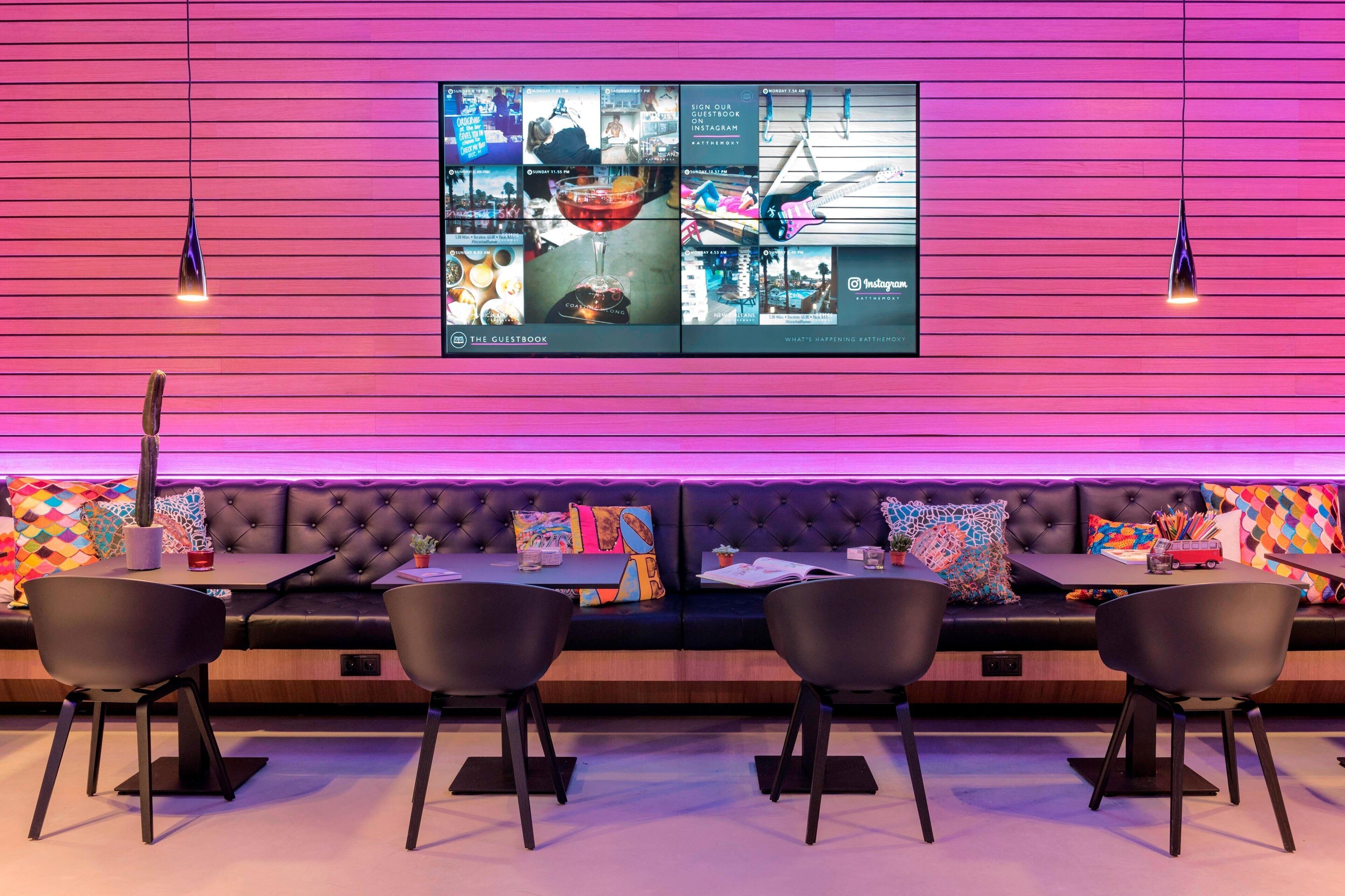 A modern lounge area with a large flat-screen TV displaying Instagram content, a tufted black sofa with colorful cushions, and black chairs around a small table.