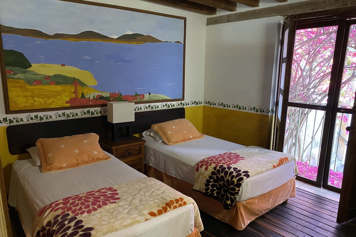 Twee enkele bedden met bloemenprint, een nachtkastje, een schilderij van een landschap, en uitzicht op roze bloemen door een glazen deur.