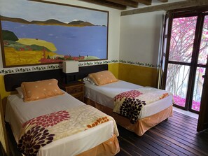 Two single beds with floral bedspreads, a nightstand, a painting of a landscape, and a view of pink flowers through a glass door. - Villa Serena Centro Histórico (Mazatlán)