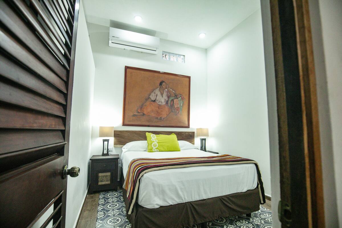 Een hotelkamer met een bed, nachtkastje, lamp, schilderij aan de muur en een vloer met een patroon.