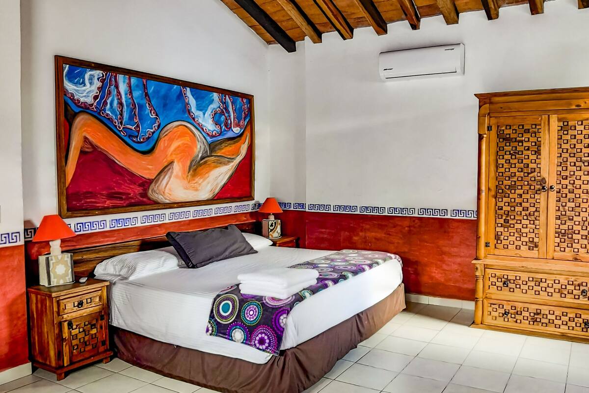 Een slaapkamer met een bed, een nachtkastje, een aan de muur hangend schilderij, een houten garderobe en een betegelde vloer.
