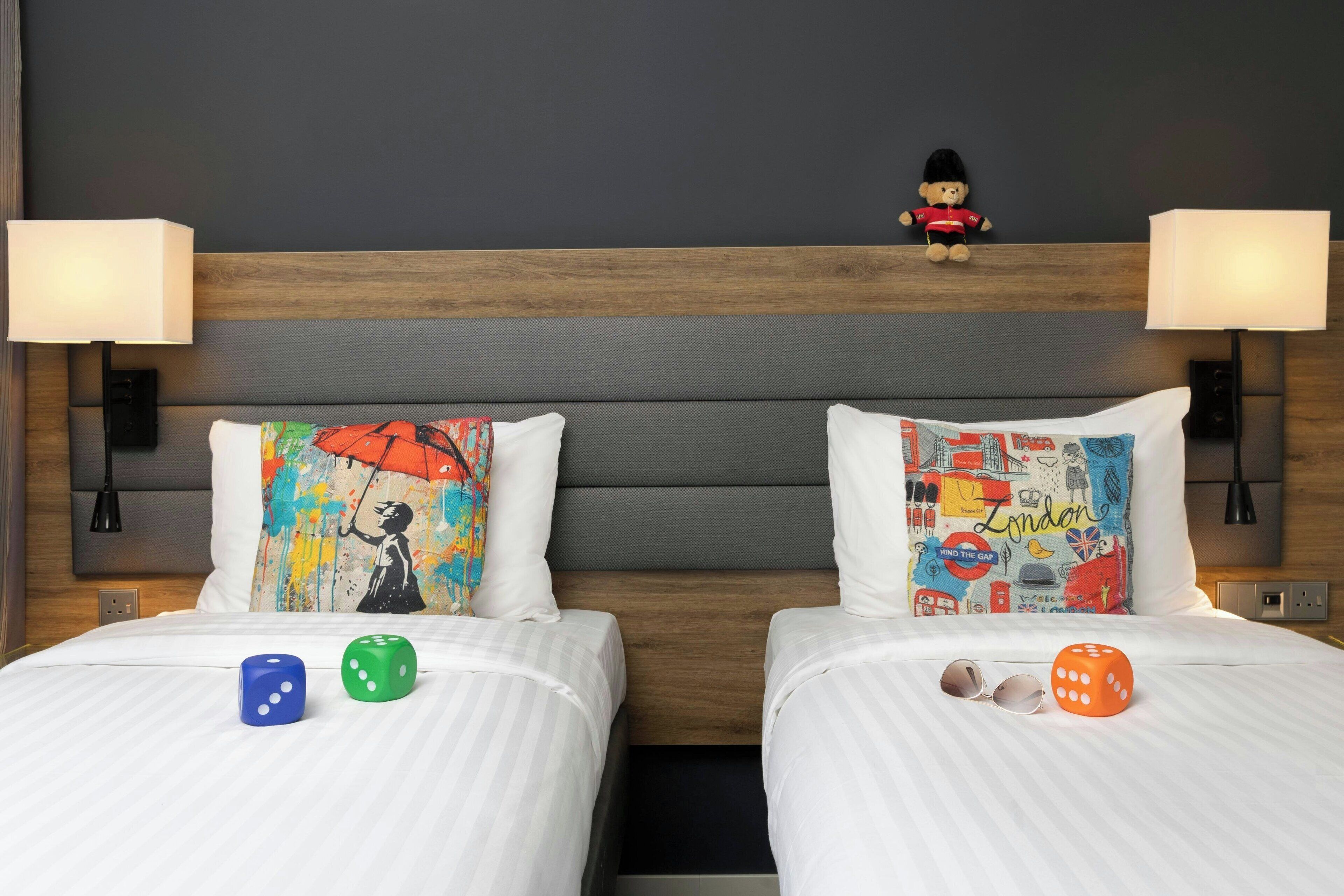 A hotel room with two beds, each with a pillow featuring a colorful design, a teddy bear on the headboard, and dice on the beds.