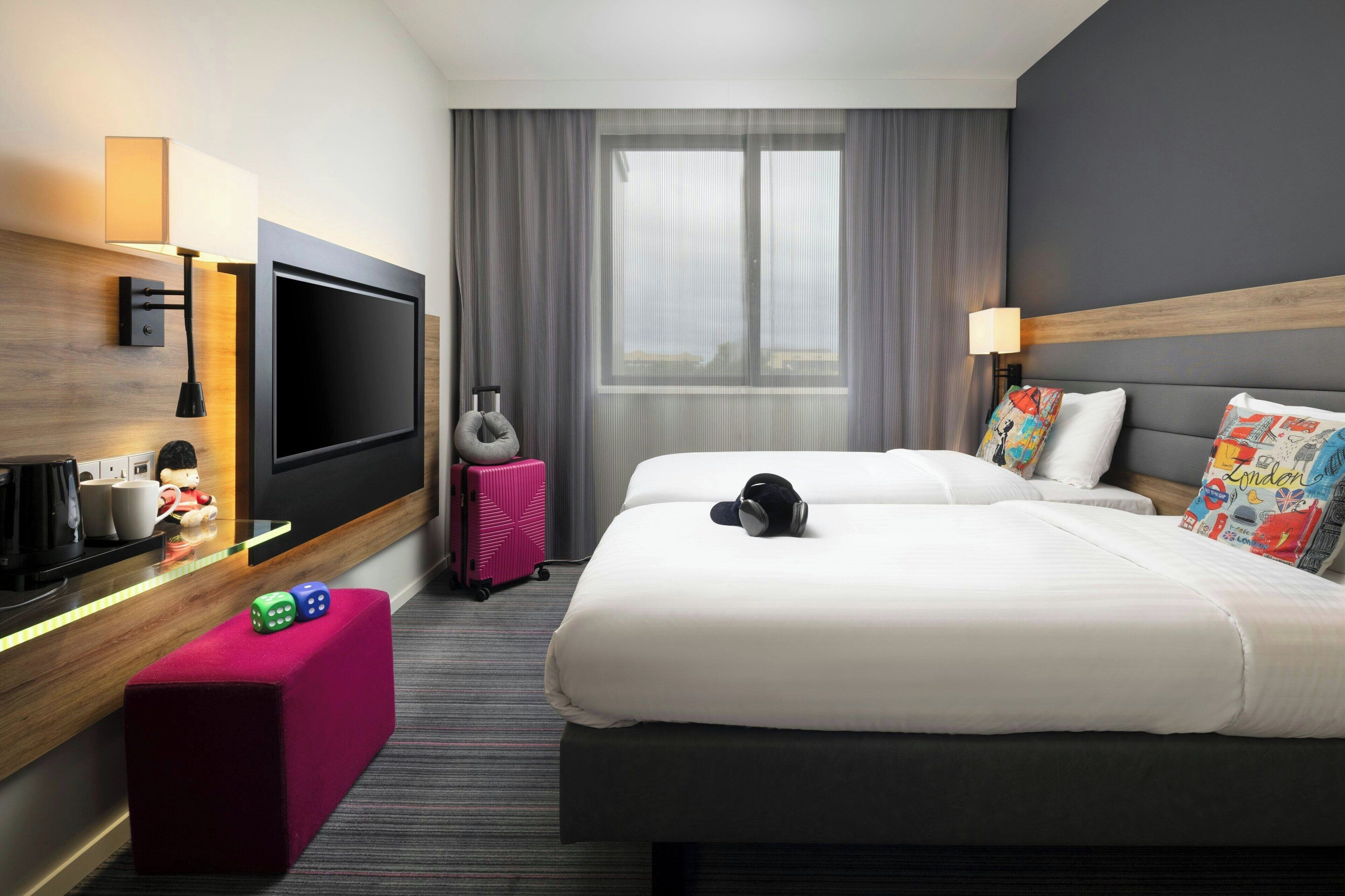 A hotel room with a large flat-screen TV, a desk with a lamp, a red ottoman, a pink suitcase, and two beds with colorful pillows.