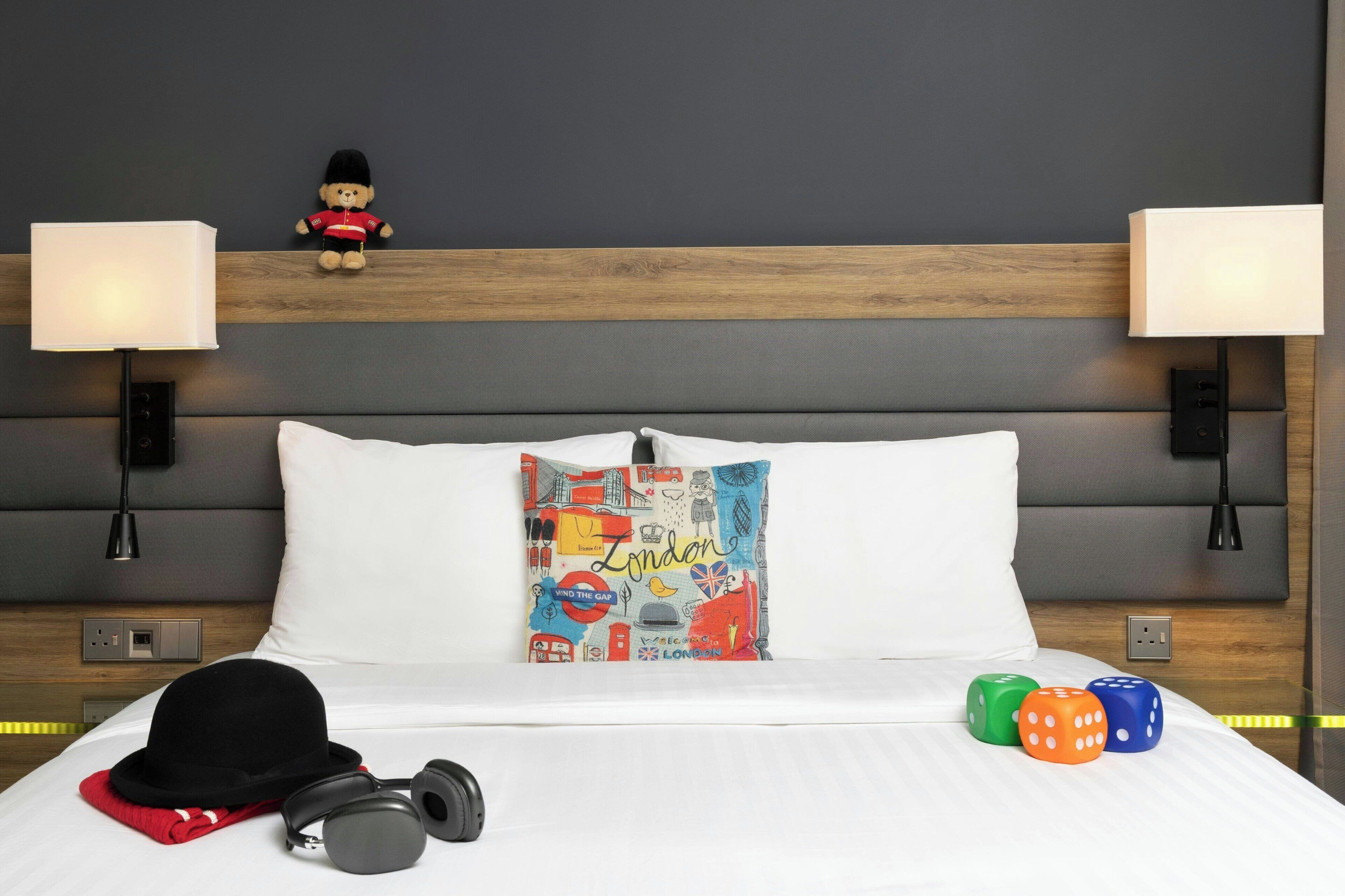 A hotel room with a neatly made bed, a decorative pillow with 'London' written on it, a teddy bear on the headboard, and dice on the bed.