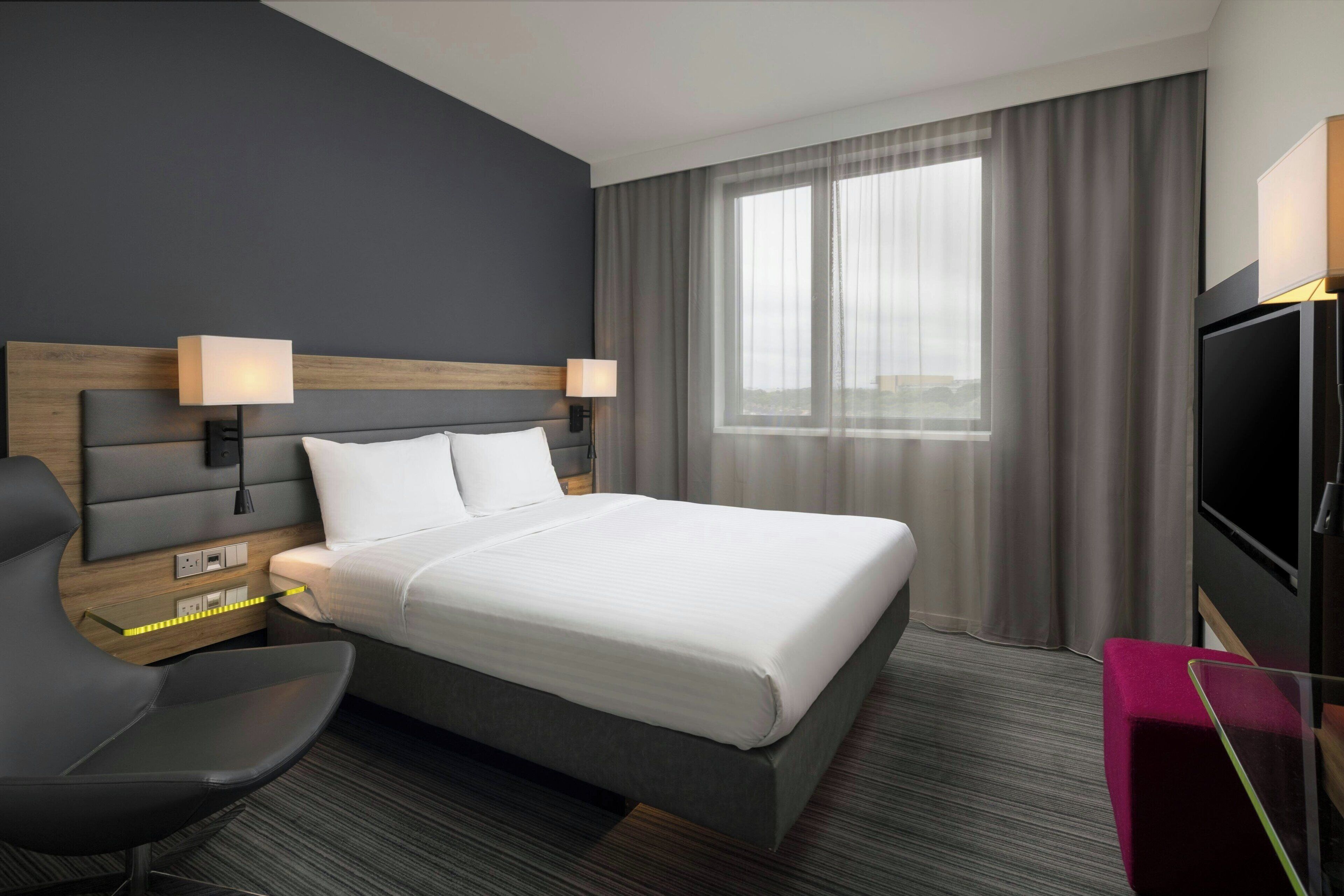 Habitación de hotel con una cama grande, dos lámparas de noche, un televisor, un escritorio con una silla y un ventanal con cortinas.