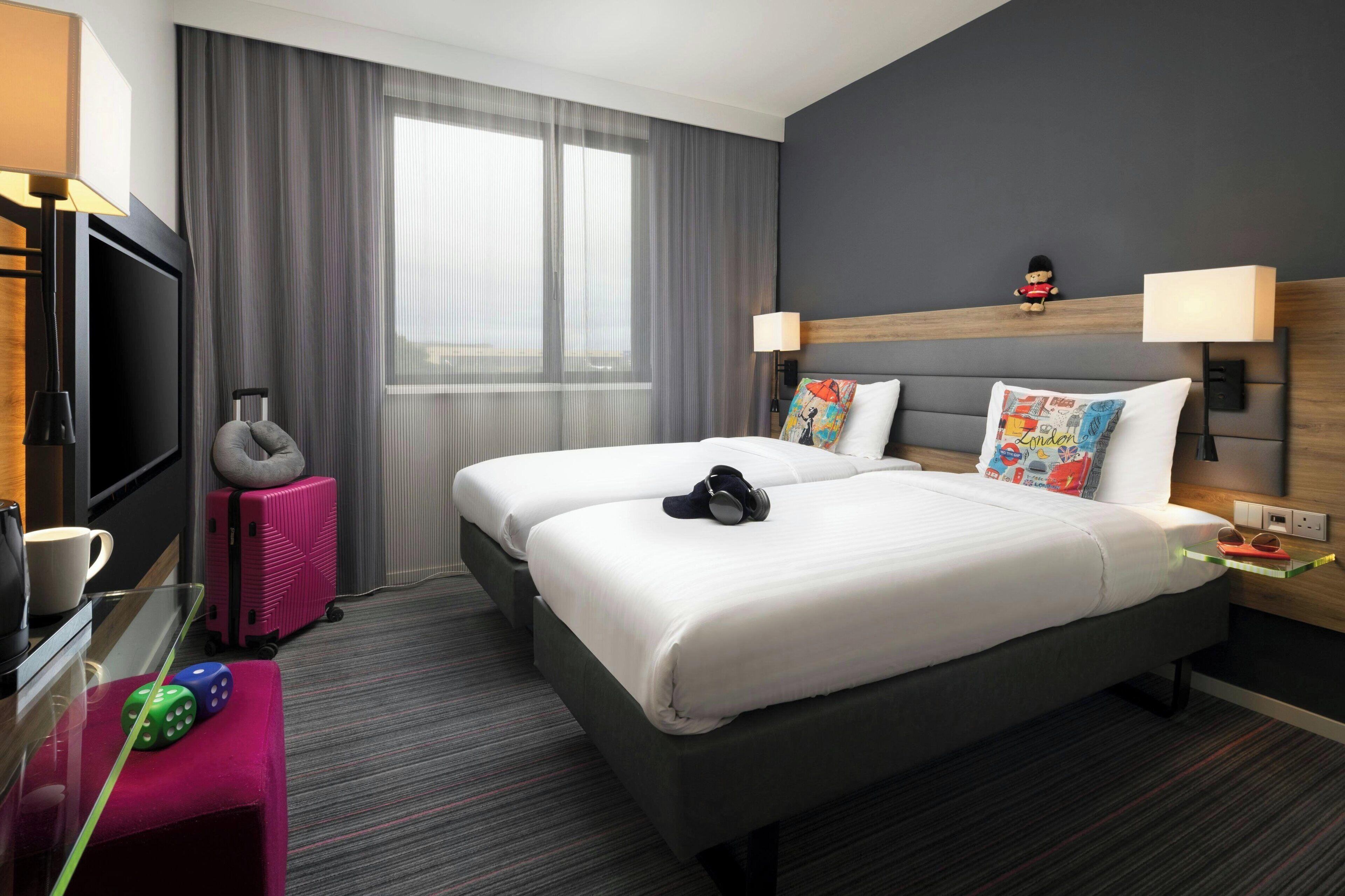 A hotel room with two beds, a television, a desk with a lamp, a suitcase, and a window with curtains.