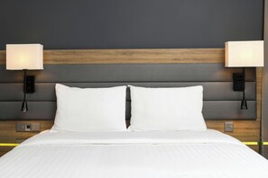A neatly made bed with white linens, a wooden headboard, and two wall-mounted lamps on either side. - MOXY London Heathrow Airport (Hounslow)